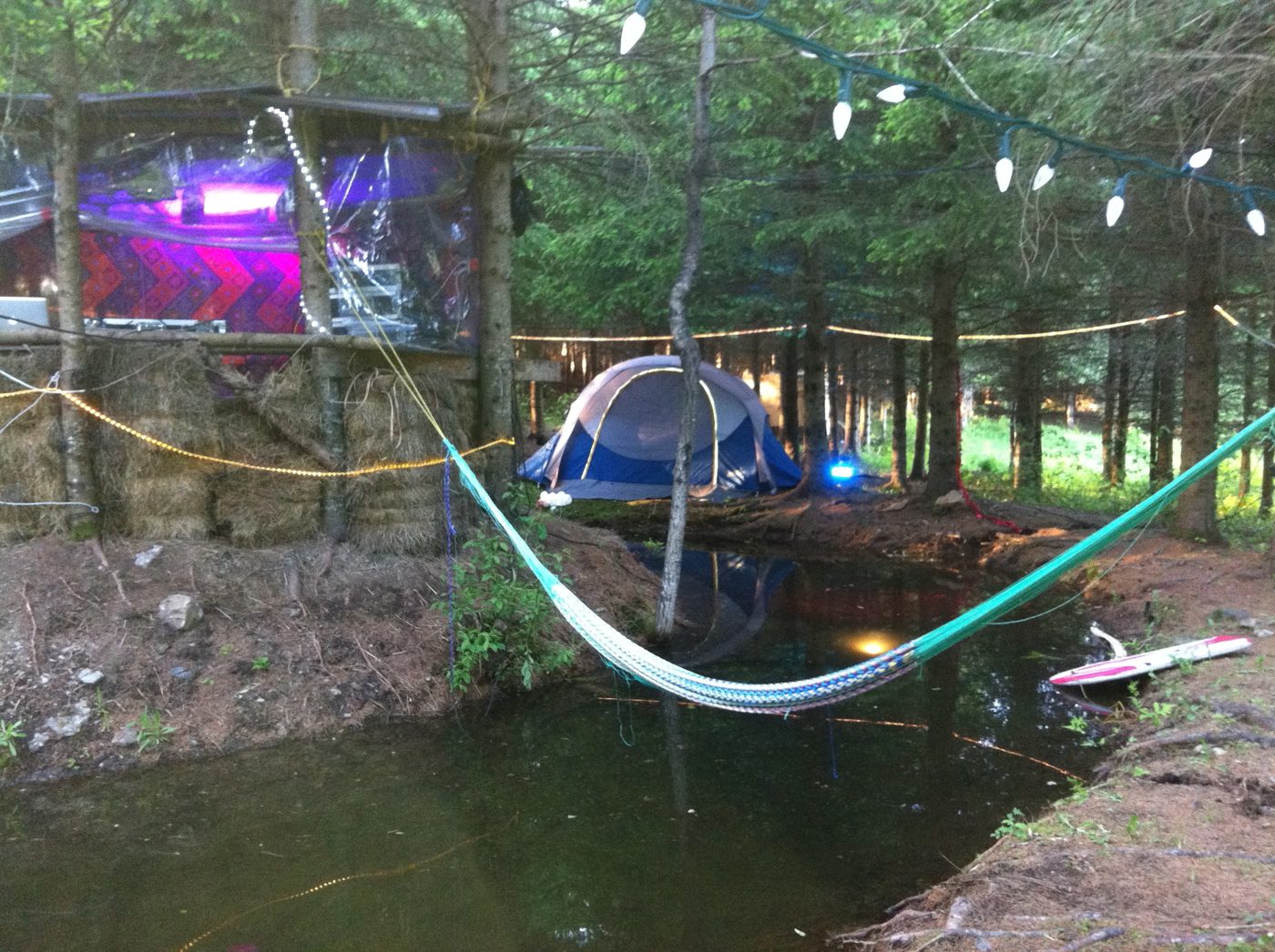 L'AgorA's Cherry Island: the cabin at the water's edge, the hammock stretched over the stream, a tent under the spruces, string lights between the trees.